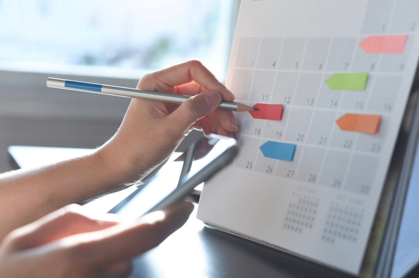 Рука отмечает дату карандашом на настольном календаре с цветными стикерами-указателями